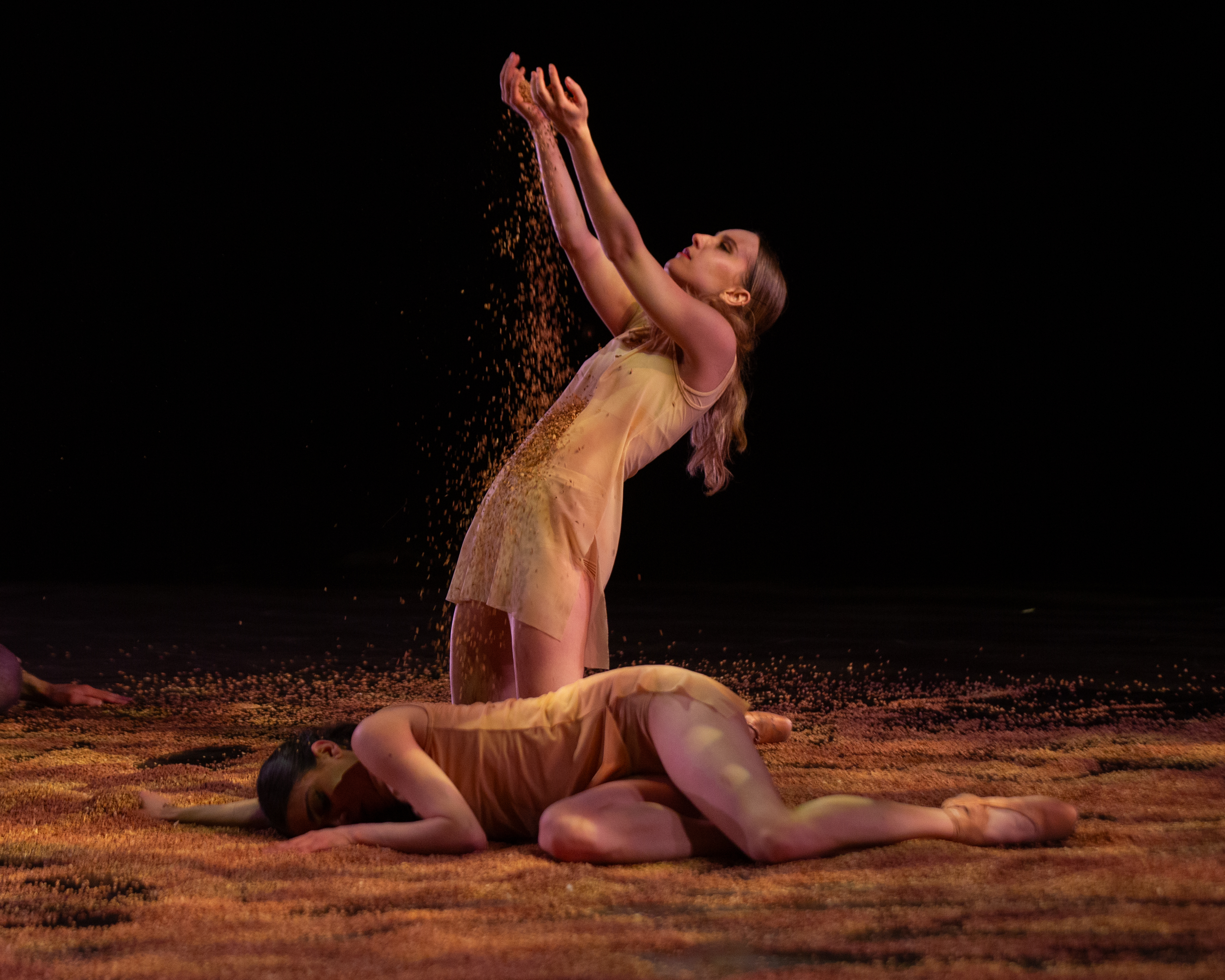 A woman dancer reaches her arms high catching shards of material. Another dancer is curled on the floor, her bare legs drawn to her..