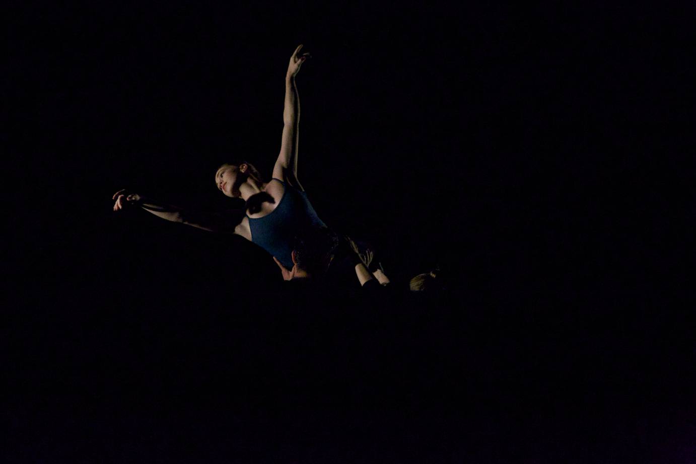 In partial darkness a women is lifted by a group of dancers in the air so that she looks like she's floating