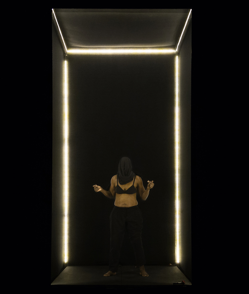 Germaine in a black bra and skirt with a black scarf over her head hiding her face while she stands in the clear box.