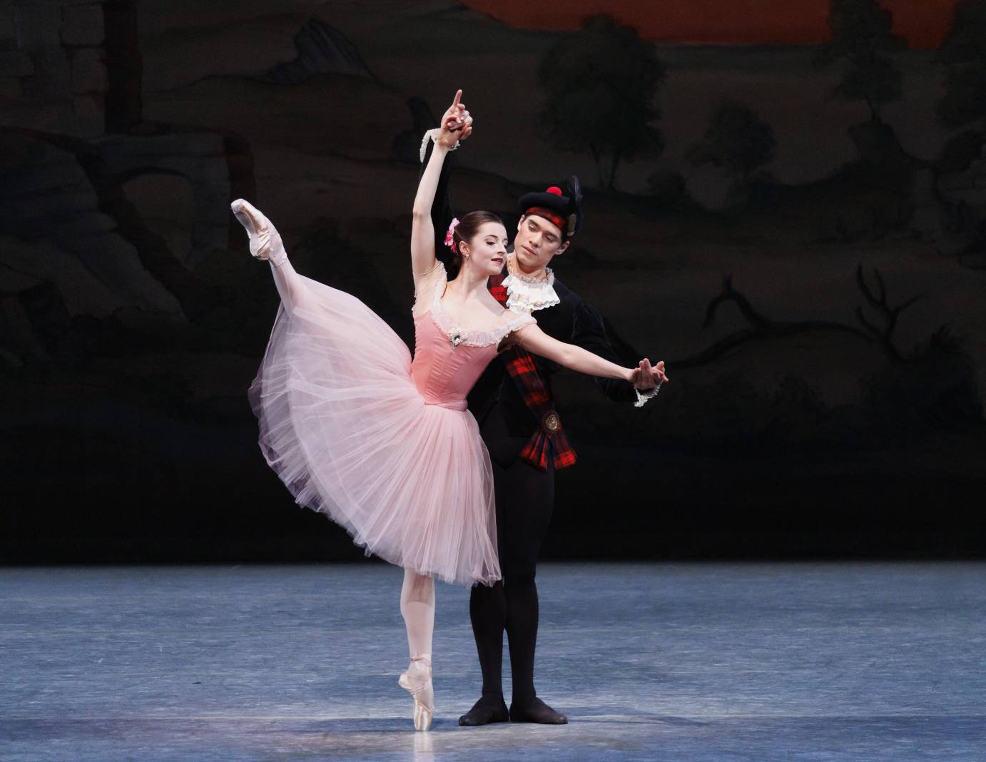 A danseur dressed in black partners a ballerina on pointe with her right leg extended in a high second position.