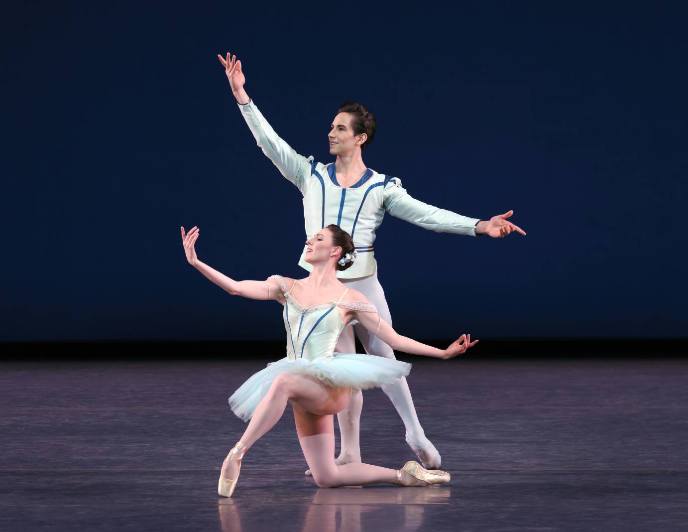 A danseur and ballerina both costumed in white with blue trim, lift their right arm while the left is horizontal. She kneels in front of him. He is standing.