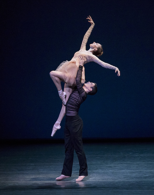 A man picks up a woman in a peach ballet costume with a flowly short tulle skirt. She looks at her outstretched hand that is directed towards the ceiling.