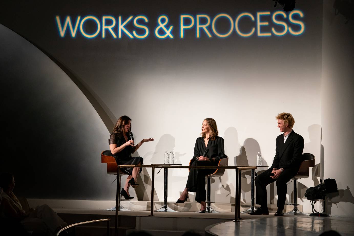 Two panelists, Kowroski and Ball, dressed in black suits, look attentively at the gesticulating moderator