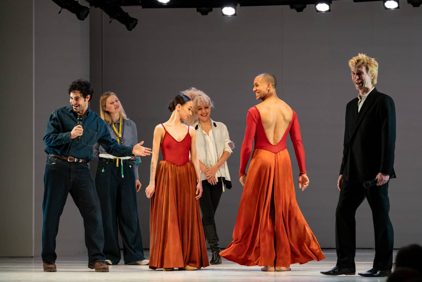 Six people, including two dancers, a female and male dressed in long red dresses, are on stage. The male dancer with back turned to the audience is dressed in a plunging back line. He smiles at the woman as she looks down over her left shoulder. Both the designer and Ball, flanking the dancers, are laughing.