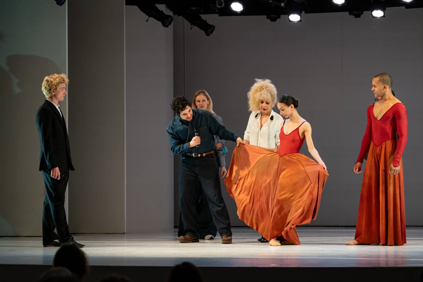 Six people, including two dancers, study the woman dancer's wide tomato-red skirt that is pulled out, presumably by the designer.