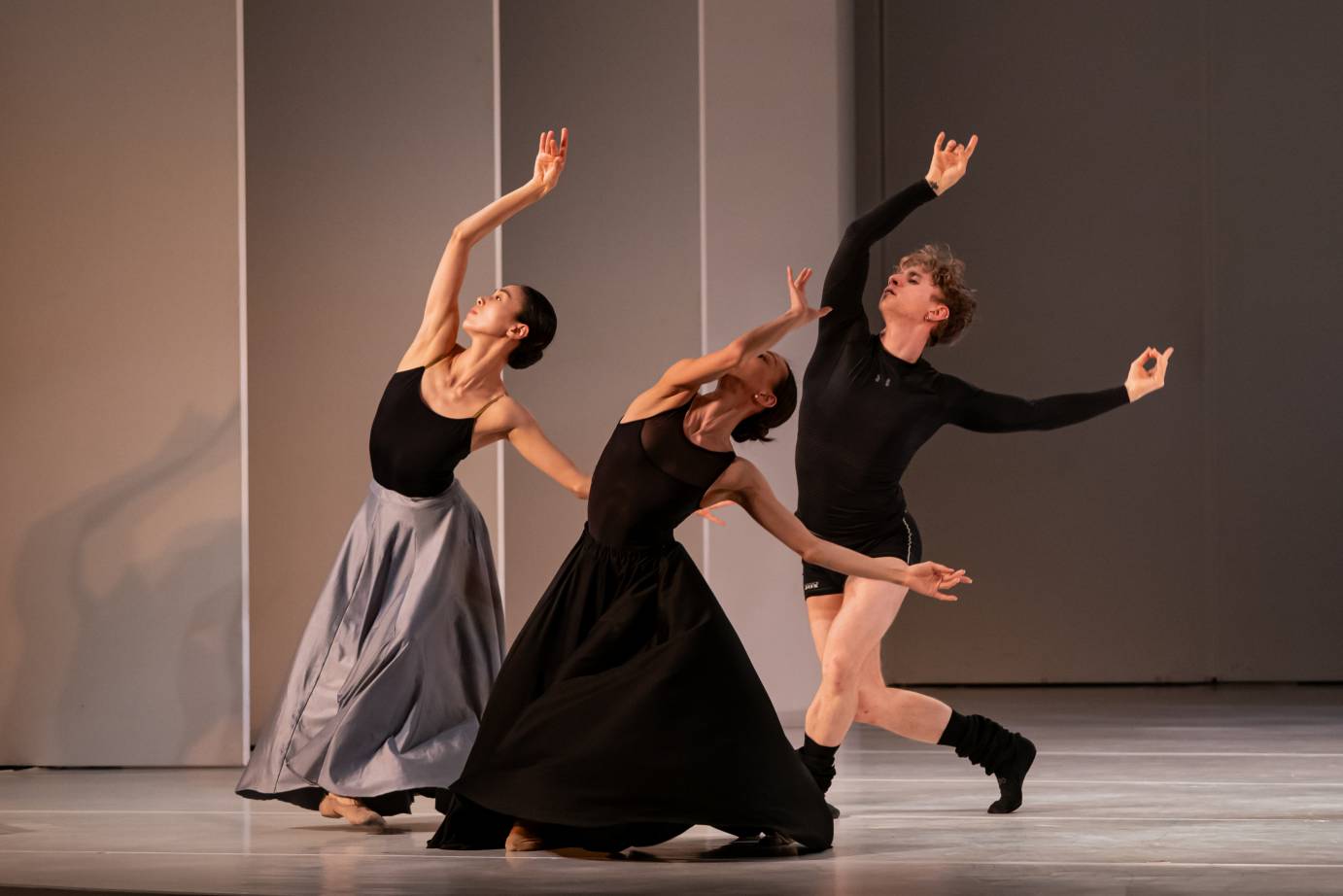 Dancers, two women dressed in long black and gray skirts and leotard tops and a man in small black shorts, a long-sleeved black top and black calf-high boots, lunge in 4th on the diagonal with right arms high, fingers gesturing.
