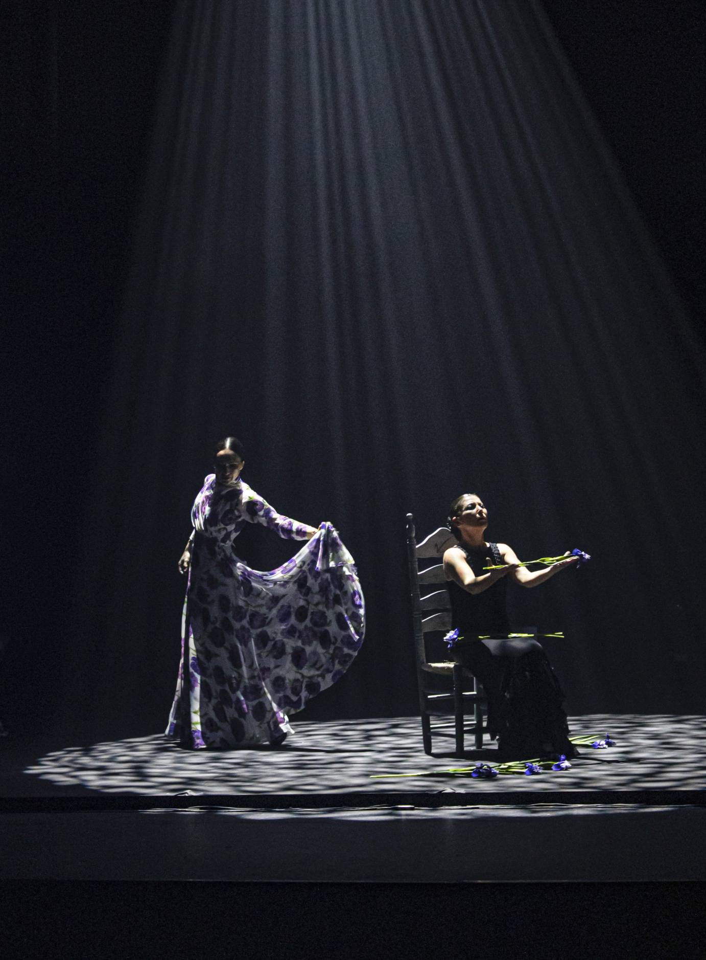 A woman in a long purple and white dress stands behind a woman seated in a chair holding a flower. The background is black.