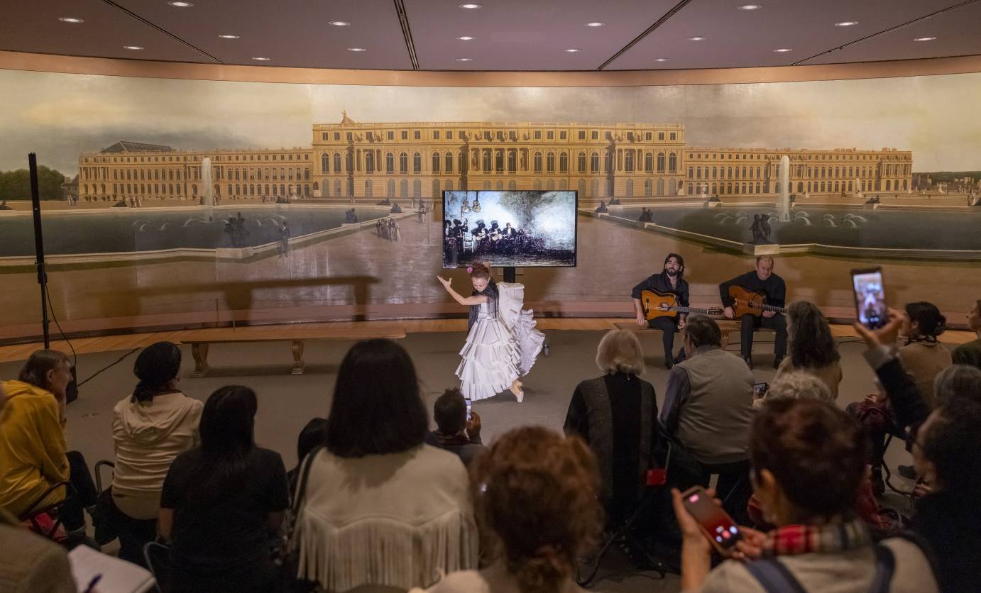A woman flamenco dancer dressed in white performs in front of a screened painting which is front of a larger painting. Two guitartists accompany.