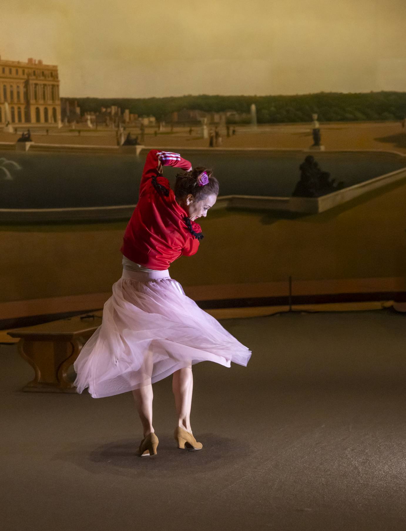 A flamenco dancer dressed in a red jacket, pink flowing skirt and high heeled shoes dances in front of a formal painting of gardens and architecture.