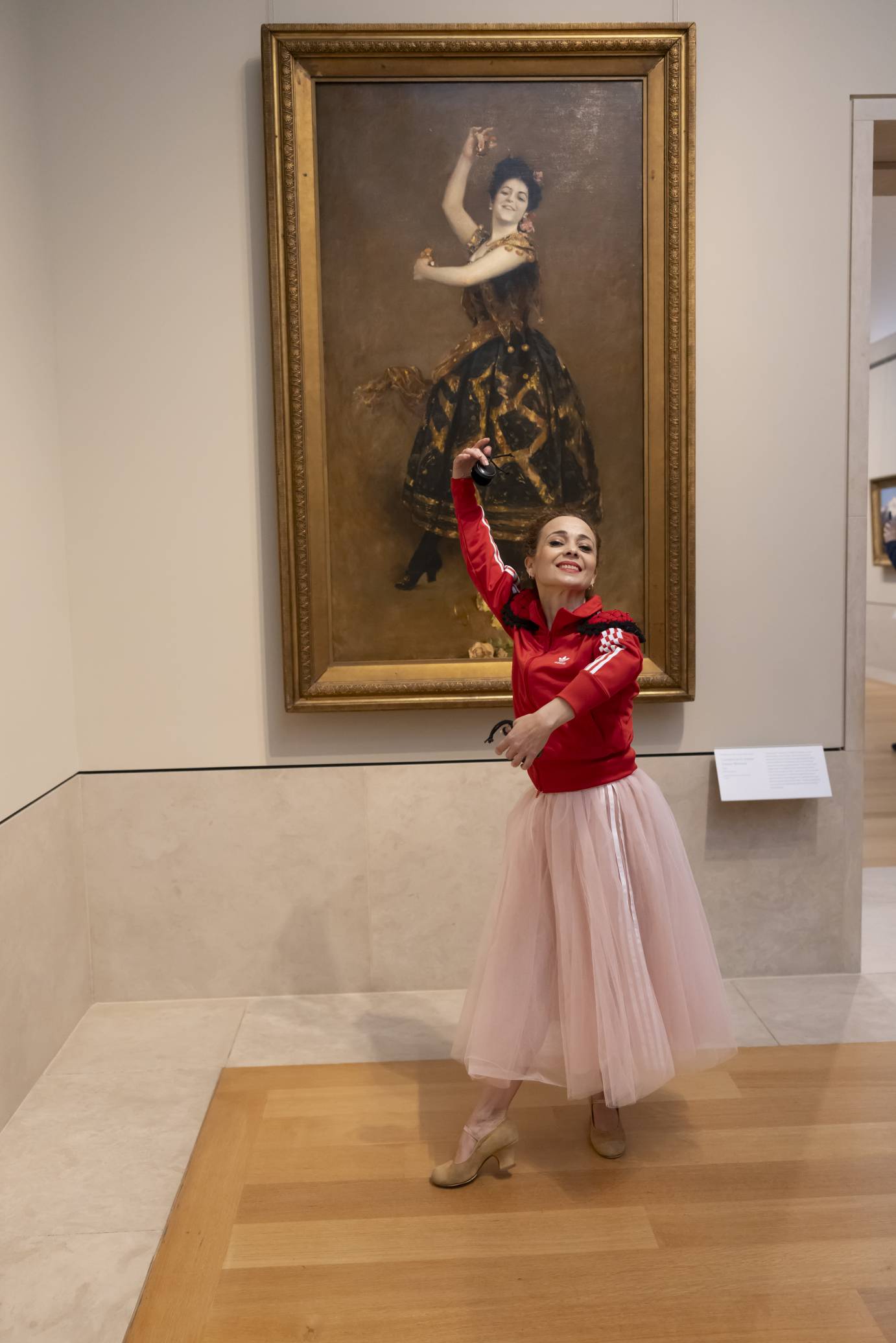 The painting of the flamenco dancer mounted on the wall behind the live flamenco dancer dressed in a red jacket and pink skirt. Their arms are in unison, each hand playing a castenet.