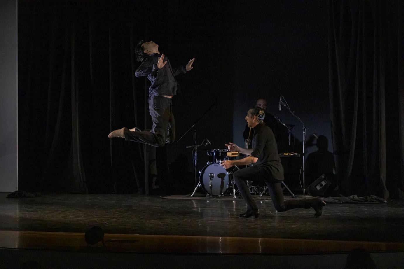 Two male dancers dressed in dark clothes against a black background. One jumps straight up with head back and palms pushing forward. The second dancer is kneeling.