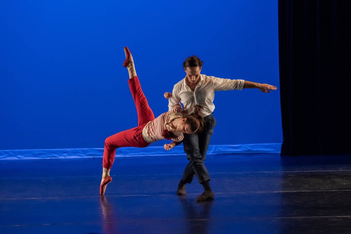 a woman in red pants and red toe shoes leans into a backwards fall supported by a man in a white shirt and pants who holds her aloft in one arm as he appears to bow in reverence.