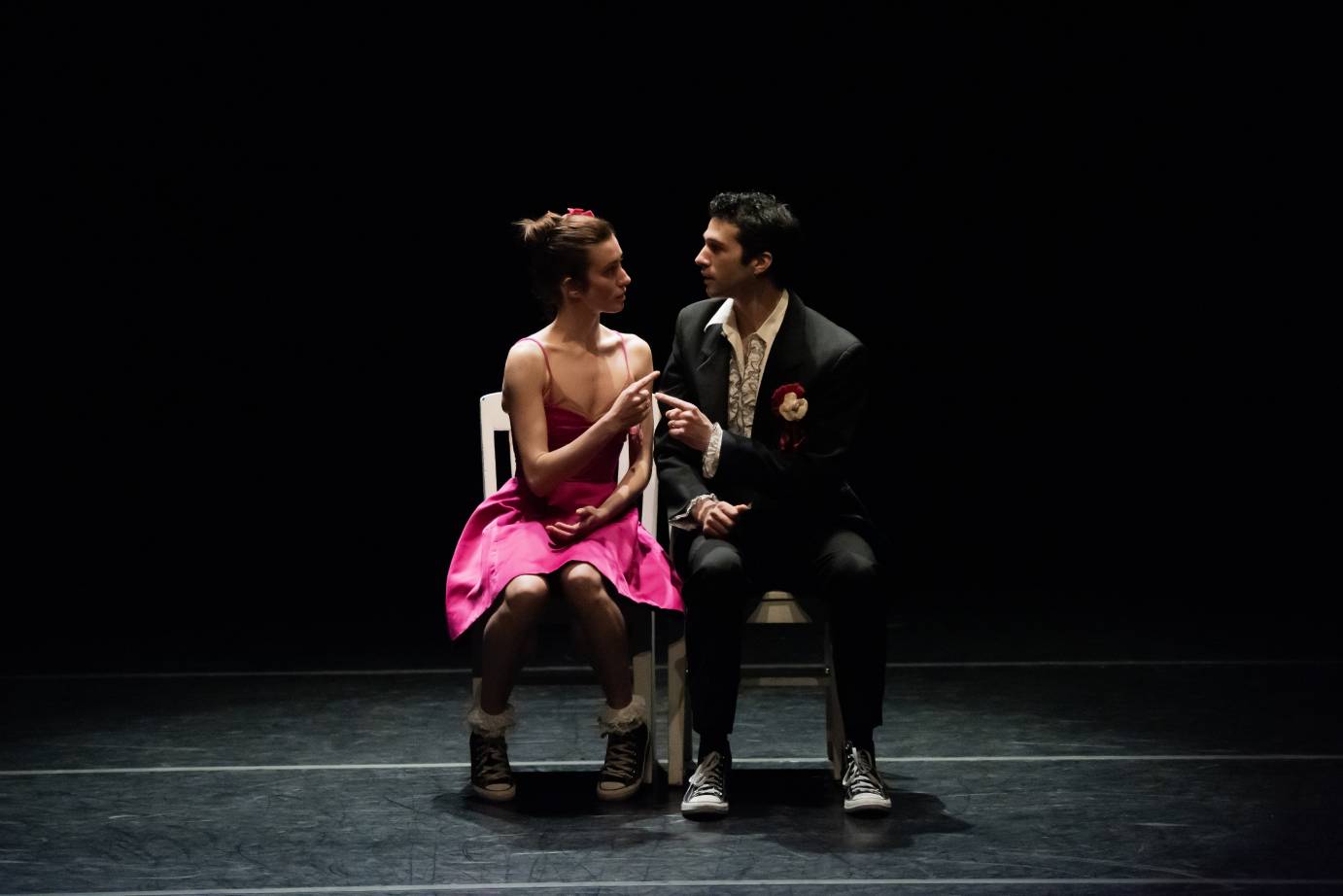 young people a gal in a pink satin dress, sneakers on her feet, a fellow in a tuxedo suit with carnation in his lapel also wearing sneakers, look at eachother quizzically. It looks like a sweet  prom night encounter. 