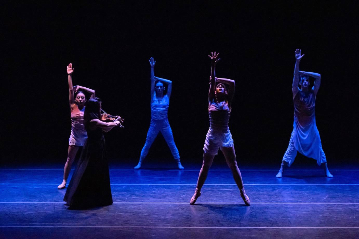 Four dancers stand held, stretched their legs are parallel and form a wide V stance one arm is raised and grabbed by the other. They seem suspended, holding their breath. A violinist in a gown walks through the space between them.