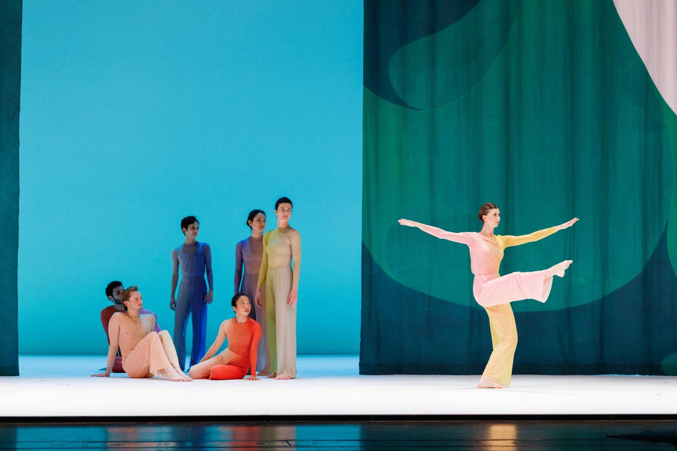  Six dancers in various colored jumpsuits both standing and seated, watch a dancer several feet away in an ochre costume her leg is extended while standing in front of a green curtain decorated with a dark green oval.
