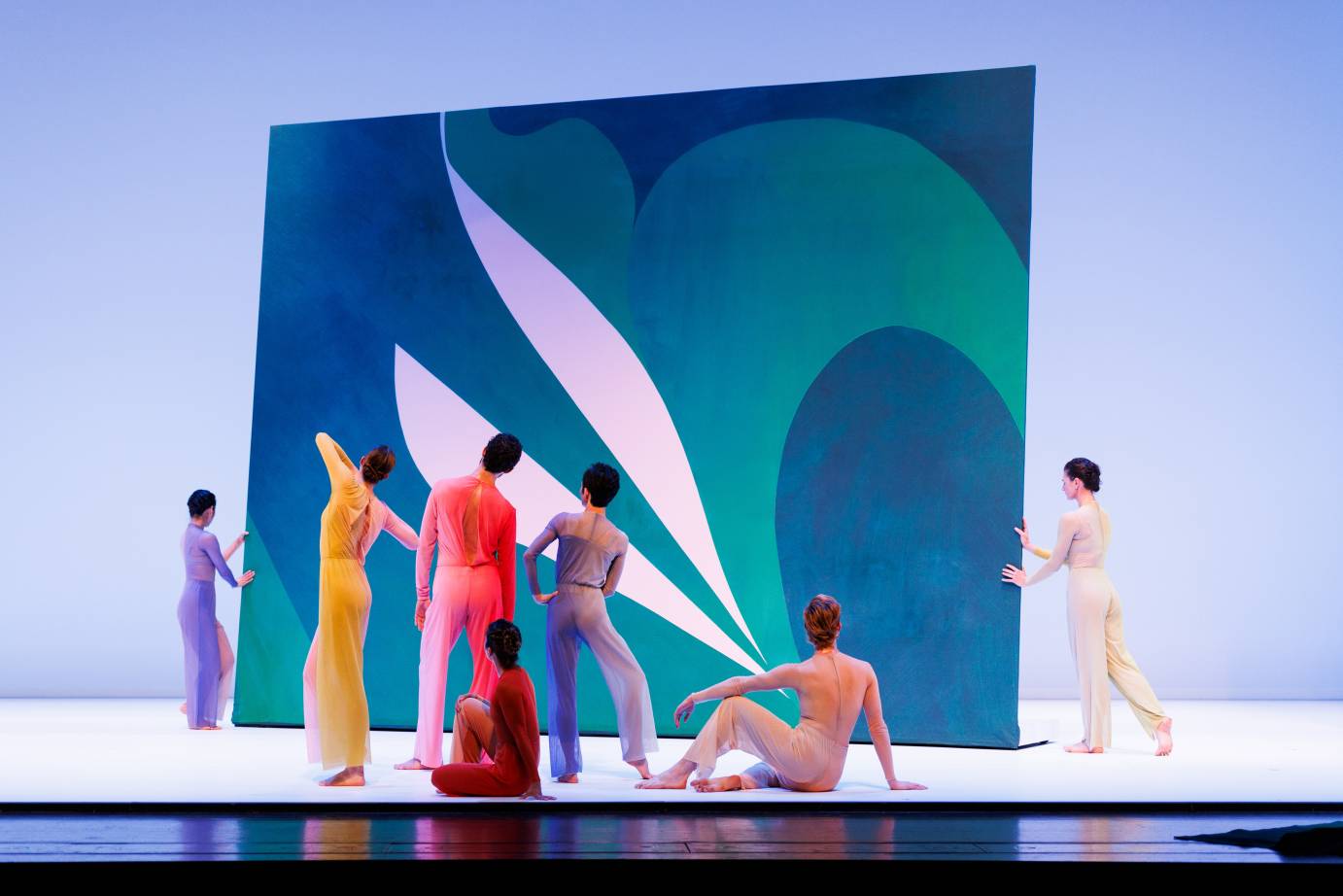 Two dancers push a screen designed with naturalistic shapes while five dancers, backs to the audience, three standing and two seated. Their jumpsuits are in colors of blue-gray, chartreuse, salmon, and ochre.