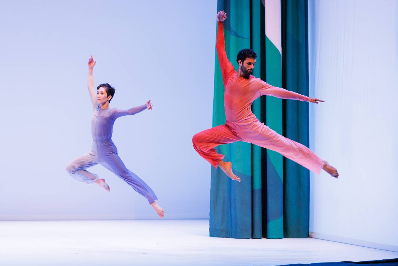 A short-haired woman in a blue grey jumpsuit leaping next to a bearded man in a salmon-colored jumpsuit. The right leg is bent while the left leg is long; the right arm is toward the ceiling next to the head while the left arm is horizontal. . The bodies face the downstage right diagonal but the woman's gaze is on the diagonal, while the man's gaze is over his left shoulder. 