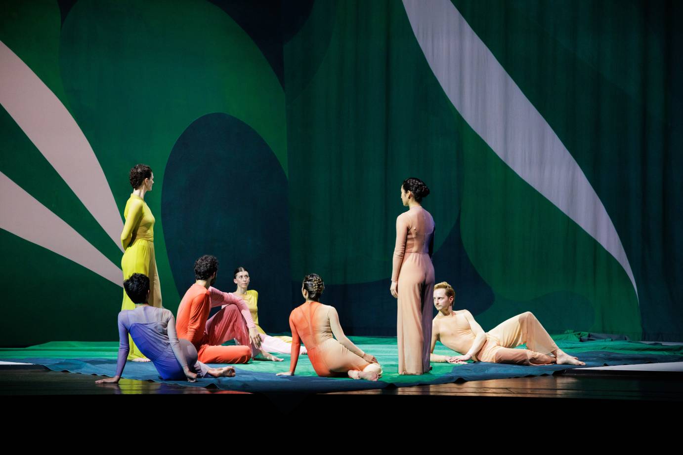 The light on stage is dark but the cast groups, five seated, legs folded to the side or knee facing up or leaning, and two standing looking at the deep green curtains decorated with naturalistic white shapes.