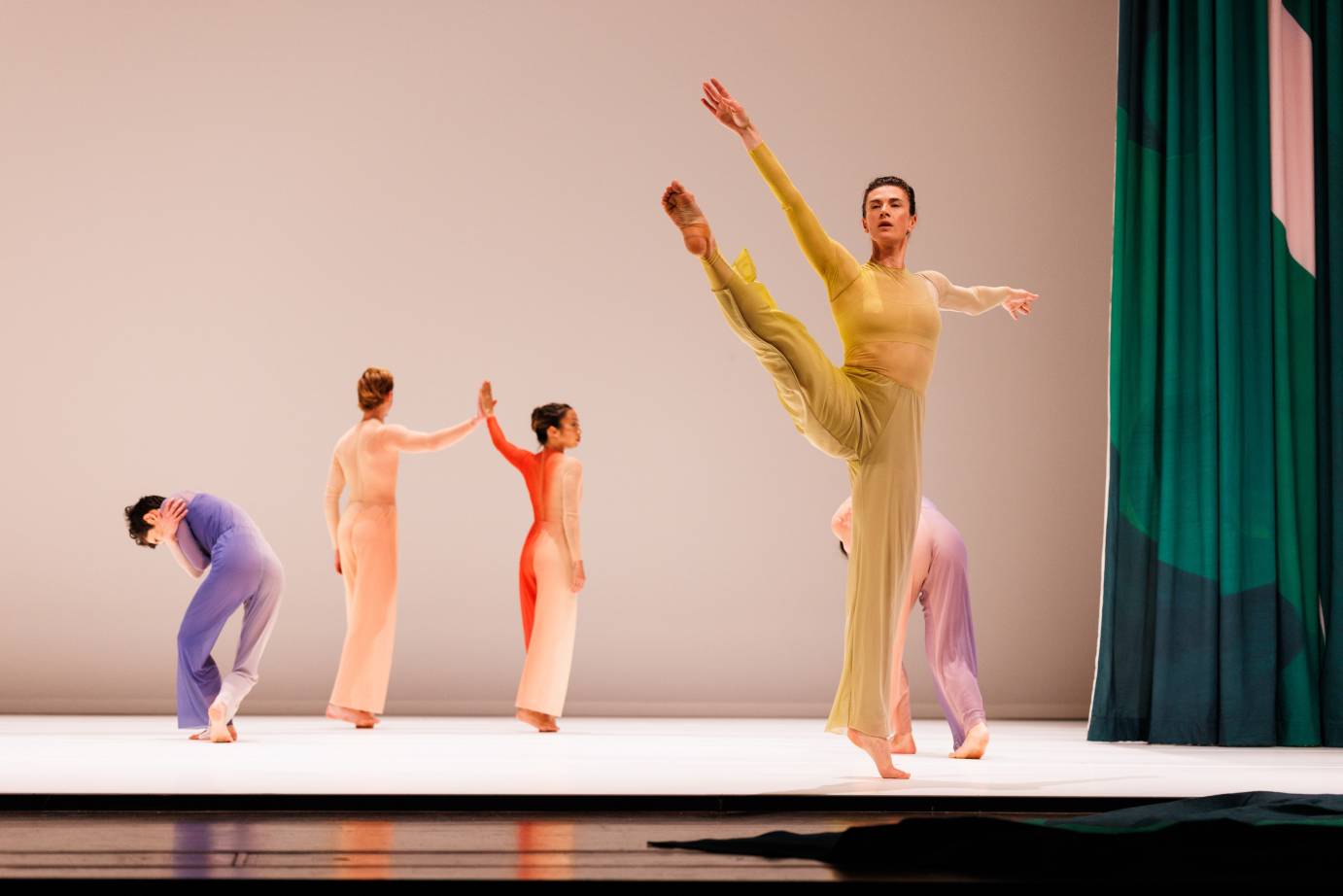 A woman on releve extends a leg on the diagonal, four dancers are upstage from her: two are touching hands and two are bent forward with arms clasping their shoulders.