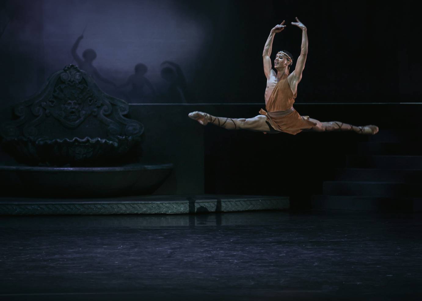 Aminta, dressed in animal-like skin over one shoulder and around his hips, leaps in a perfect horizontal split with arms extended above his head. An expression of ease on his face.