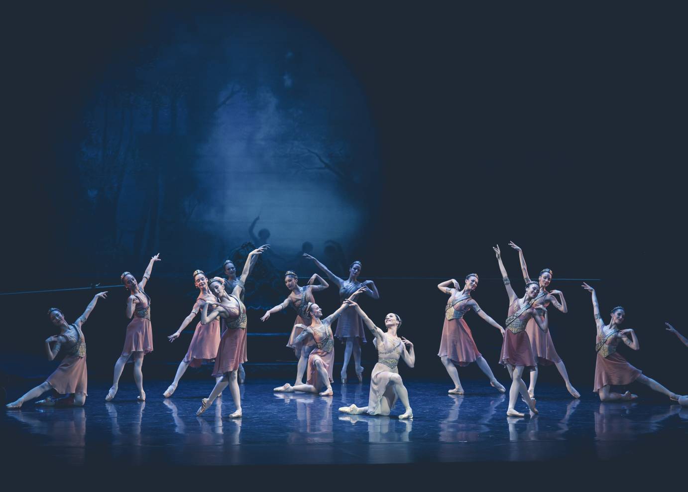 An ensemble of huntresses in rose-colored skirts and gold-flecked bodices forming a semi-circle around the primary dancer, in light pink, on bended knee. Arms are lifted to the ceiling or down on the diagonal. Fingers of the other hand rest on a shoulder.d-