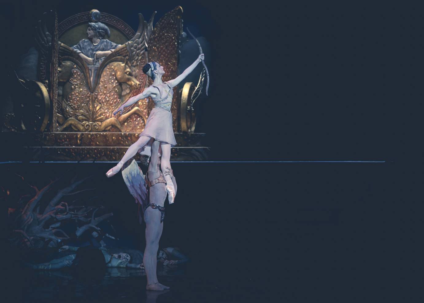 A winged Eros in a dance belt lifts Sylvia, in pink dress carrying a bow, above his head against a gold-sculpted backdrop.