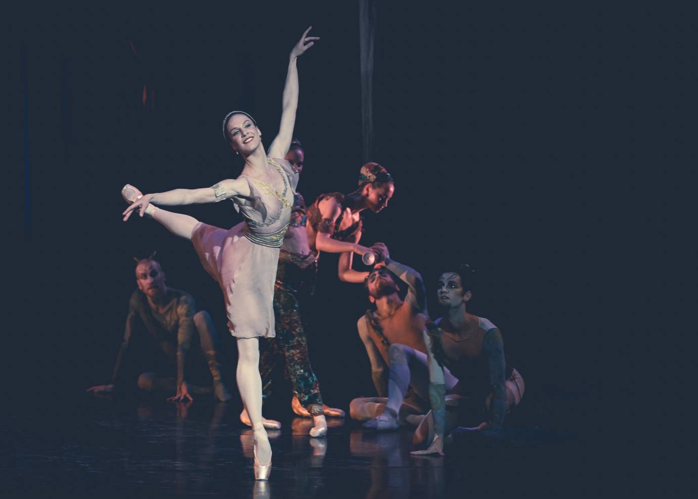 Sylvia, in a pink dress, radiates joy while perched in back arabesque, one arm above and the other arm horizontal.