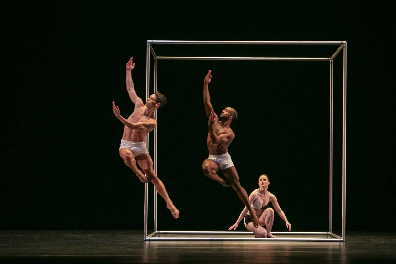 A woman kneels in the cube set piece looking up at two leaping men in light shorts, chests bare, one leg long, the other in parallel passe, one arm long, and one arm across the body. They move in front of the cube.