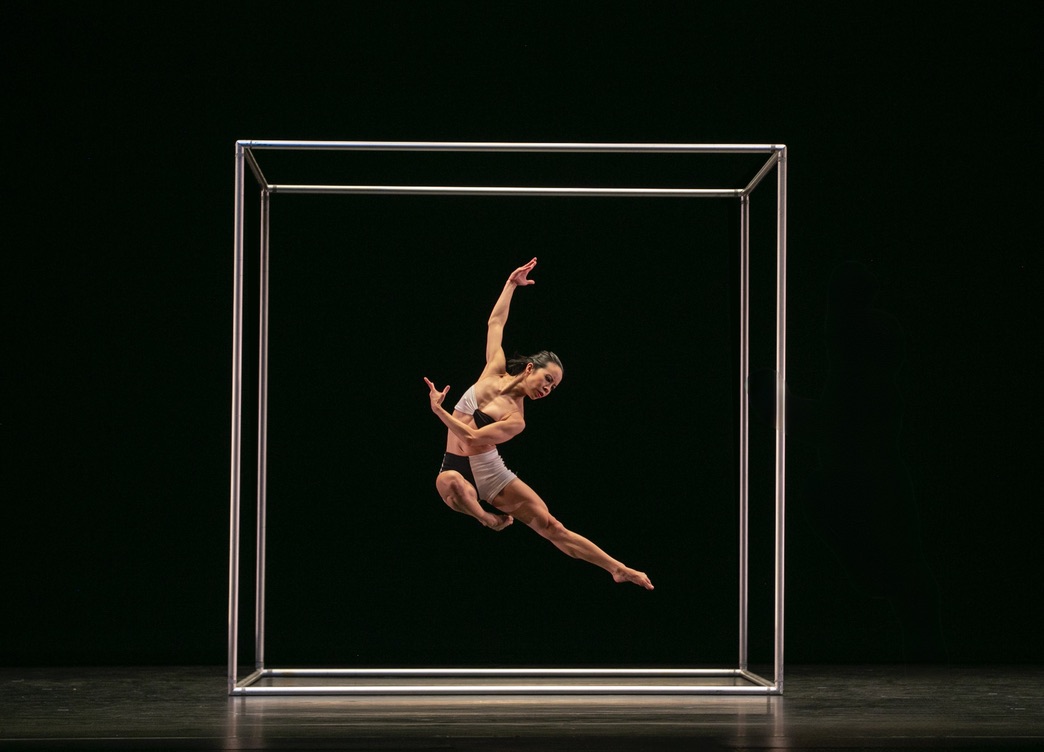 A woman leaps with body curved, one arm to the ceiling and the other crossing her body, one leg long, the other in parallel passe. She is framed in a cube set piece.