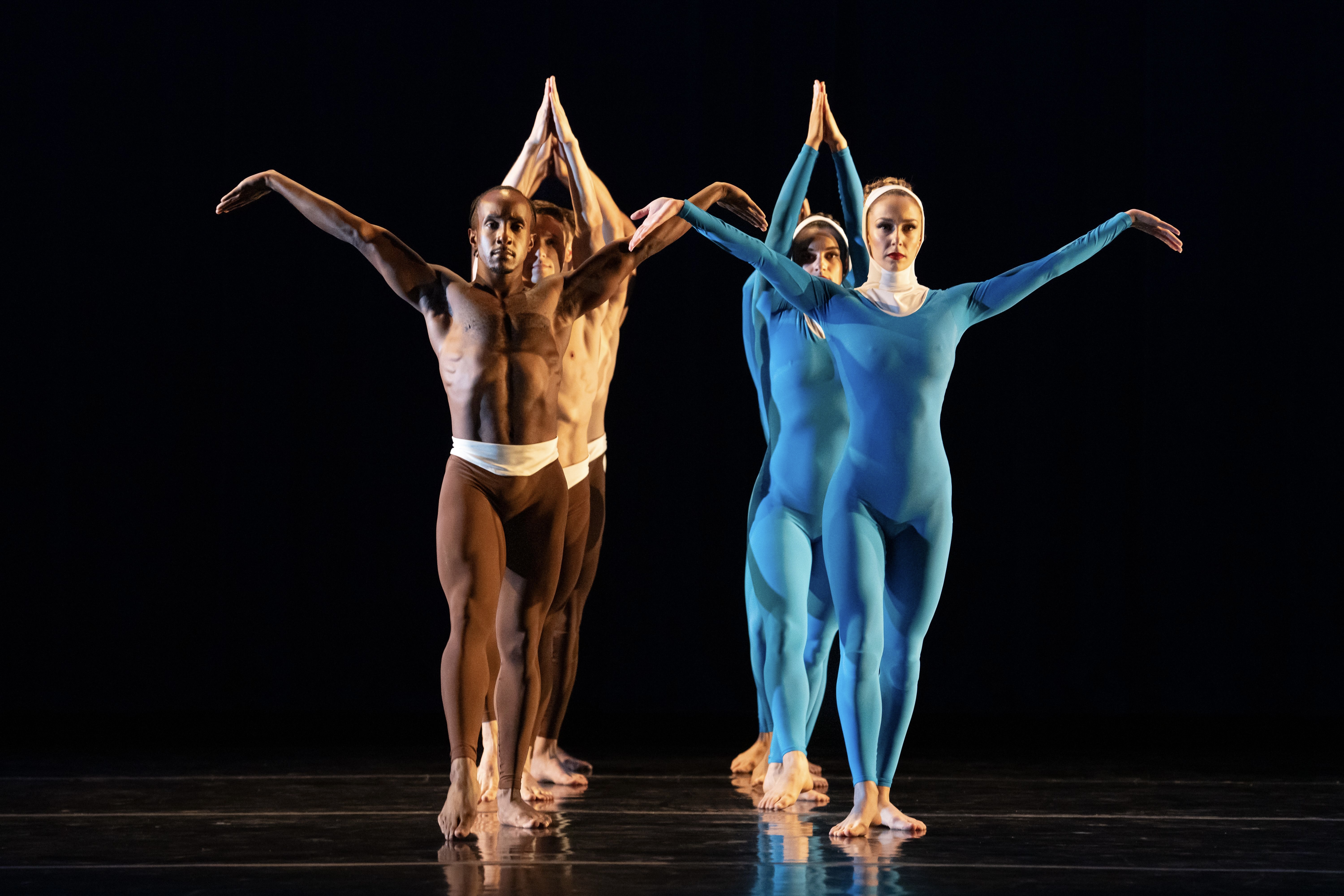 Two men on the left, barechested in brown unitards and cream-colored cumberbunds, and two women on the right, in blue long-sleeved unitards, align two front two behind facing downstage. The man and woman in the front open their arms in an upward V, wrists flexed, palms outward.