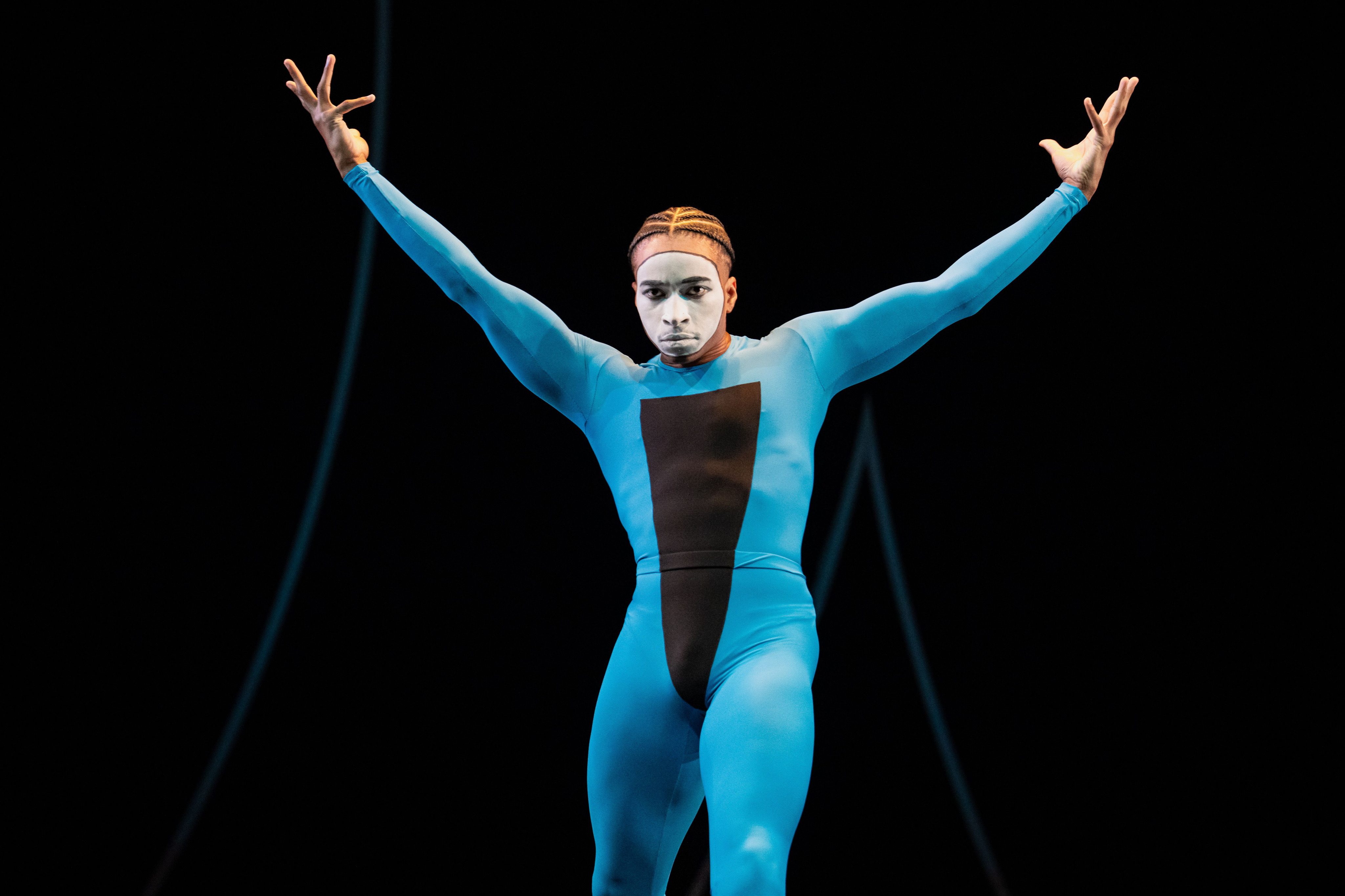 A man whose face is ovaled in white, in blue unitard with a long, black triangle pointing to his genitals, lifts his arms on the diagonal while looking forward.