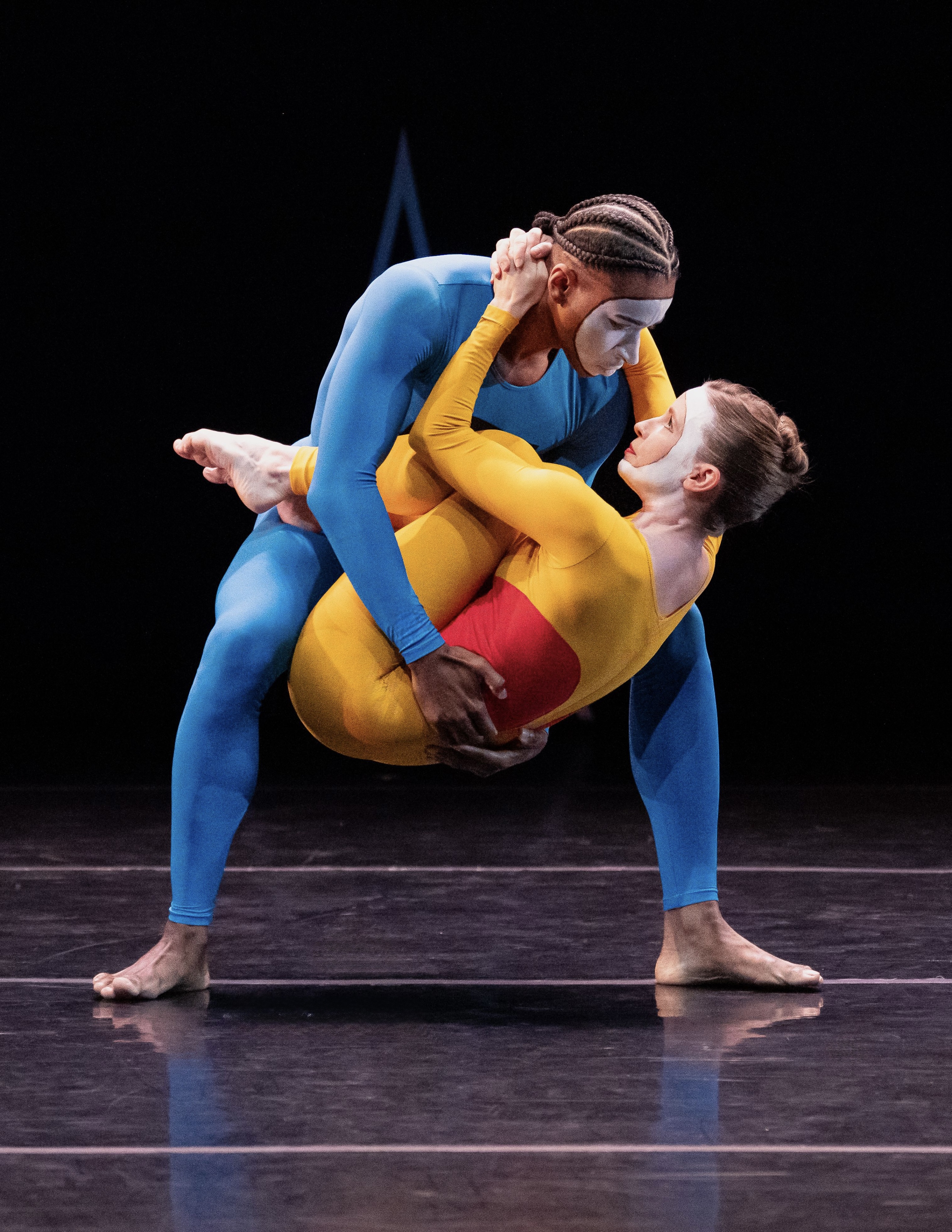 A man in blue cradles a woman in yellow, both with white on their faces, looking into each other's eyes.