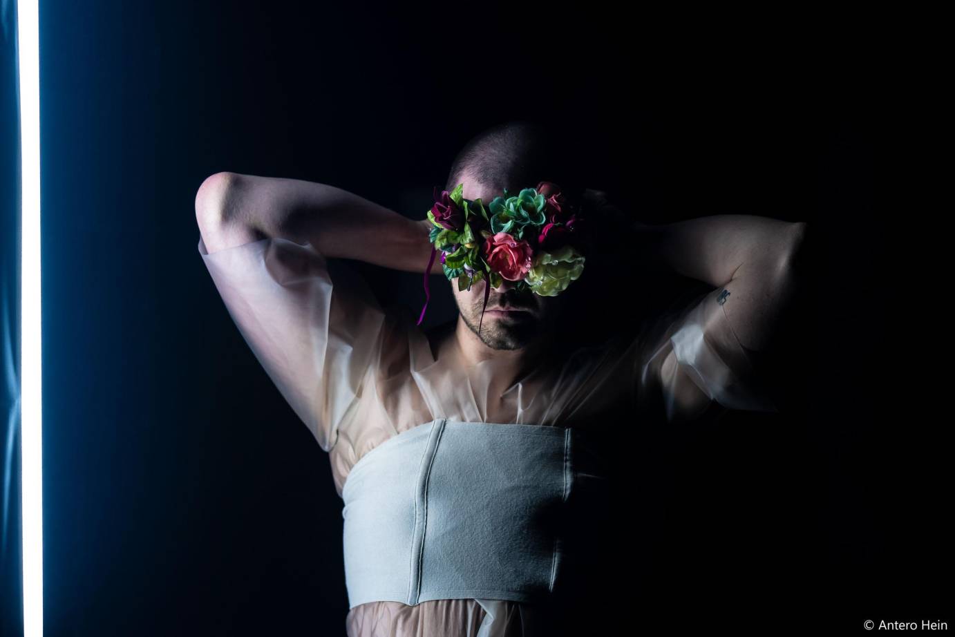 bearded man in sheer balletic looking top facing front arms up with flowers covering his eyes