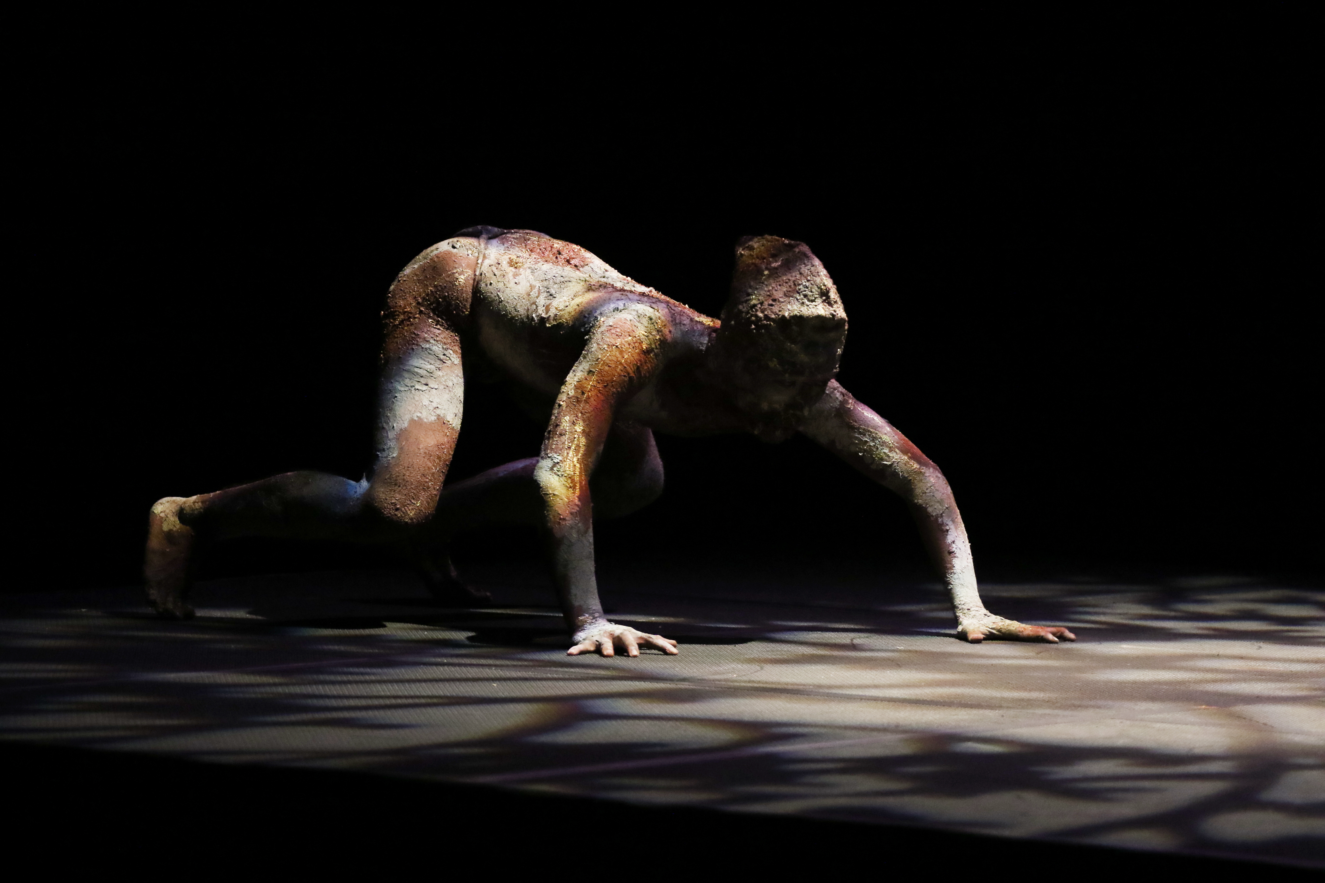 A dancer creature whose costume is made of mud in various earthen colors scampers on all fours with their pelvis higher than their shoulders.