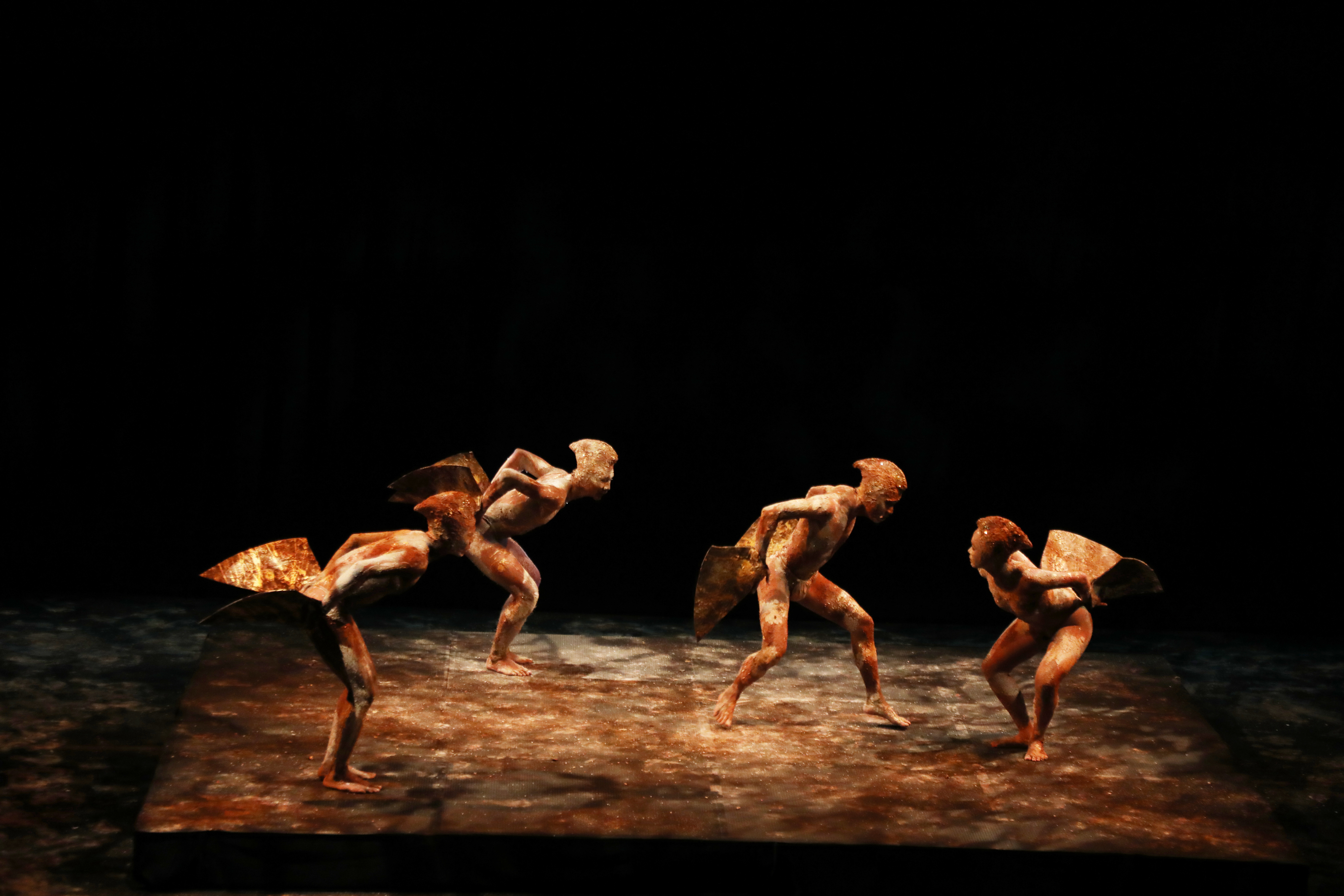 Four bird-like creatures, evenly spaced, traverse in a circle, their bodies bent forward, arms behind them with elbows bent.