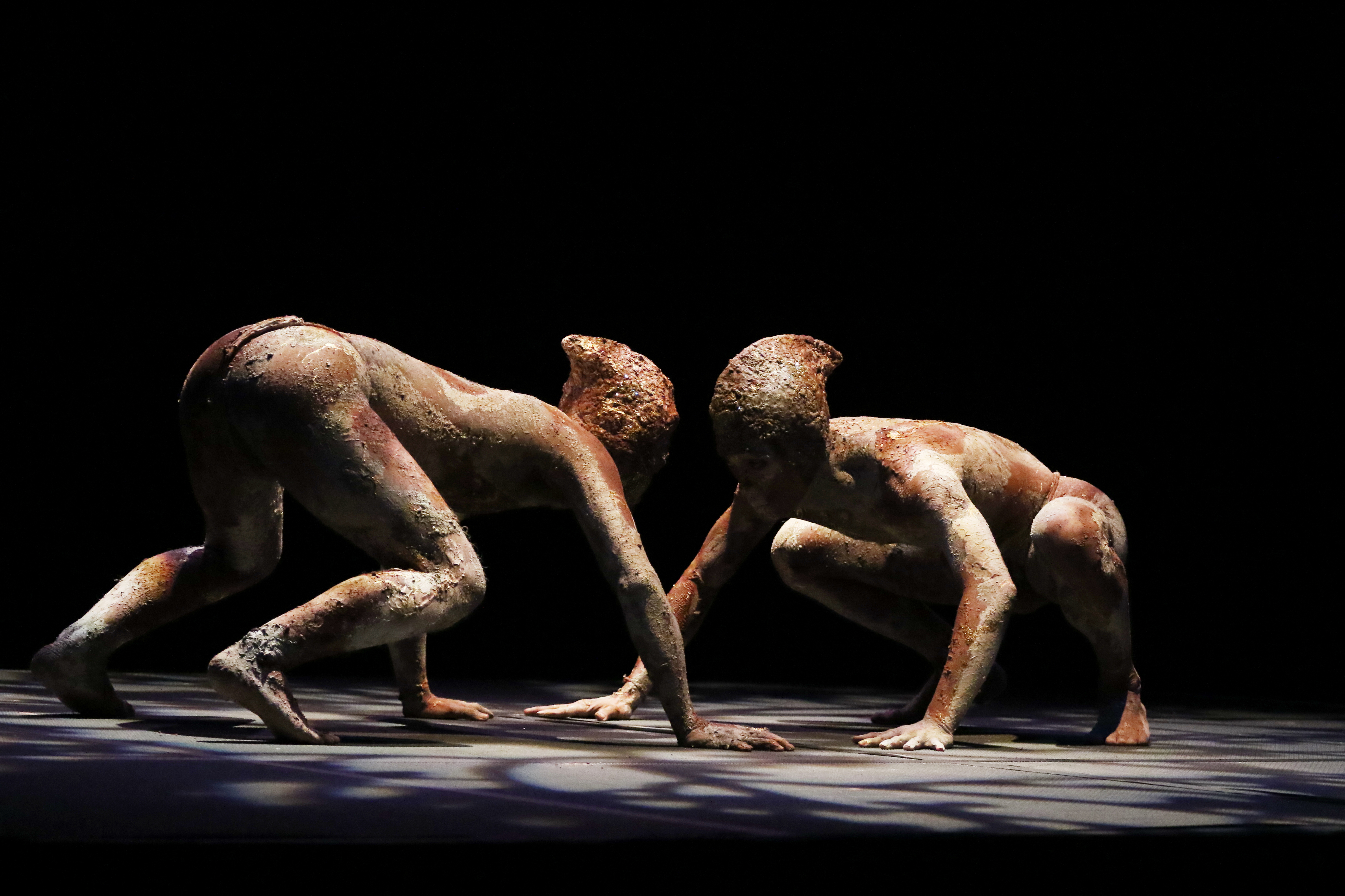 Two mud-creatures face one another on all fours. The pelvis of the creature on the left is high, while the other creature's pelvis lowers to the heels. Their heads are close.