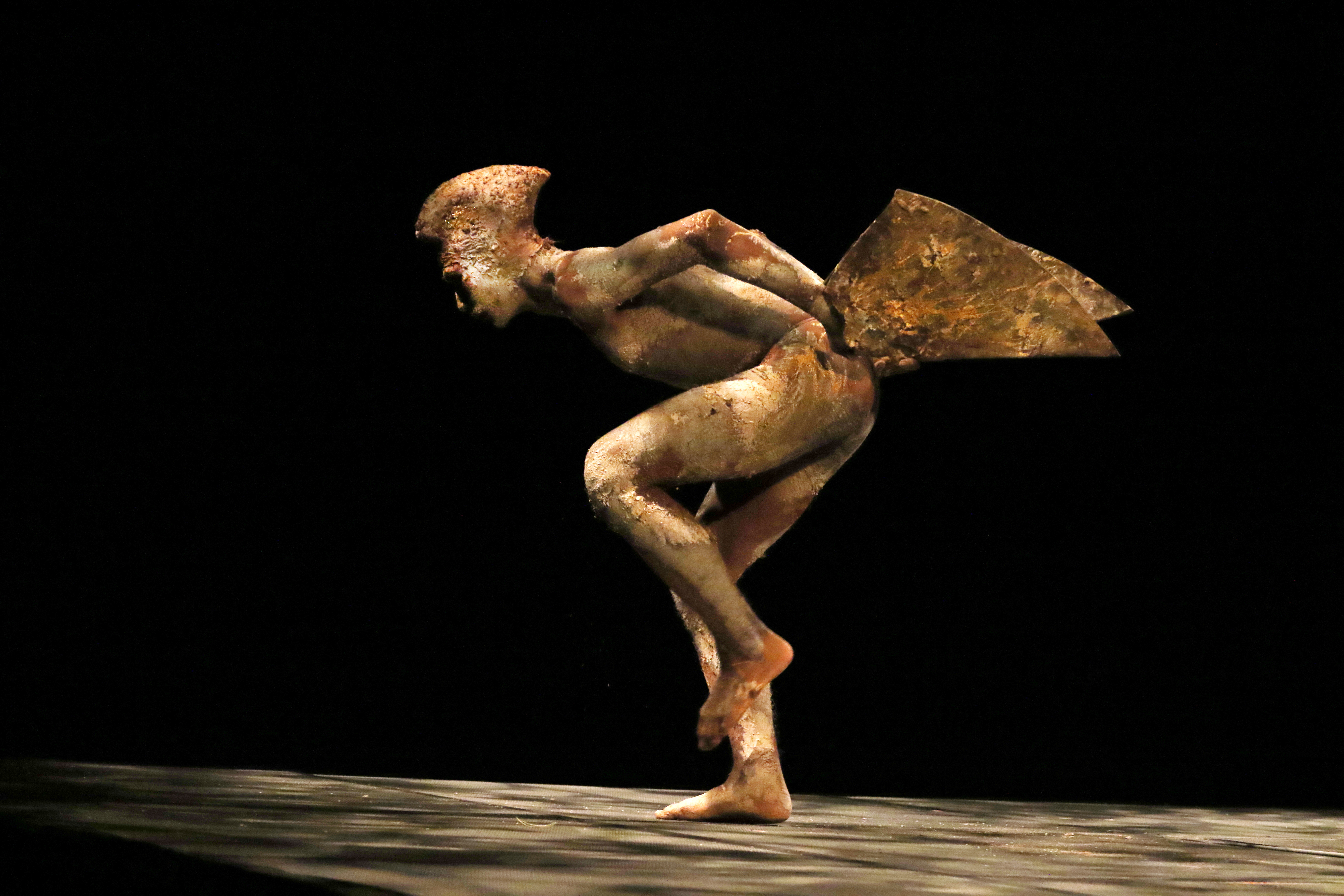 A golden mud-painted dancer depicting a birds dons wings jutting from the pelvis and a headdress that seemlessly grows from the head. The figure faces sideways, folds from the waist while standing on one leg. The other leg is slightly bent at the knee