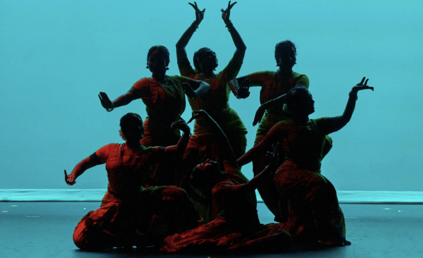 A darkened group of dancers are backlit by a luminous blue torquoise. Their poses are on various levels while their arms and hands are overhead, infront and to the sides of their bodies.