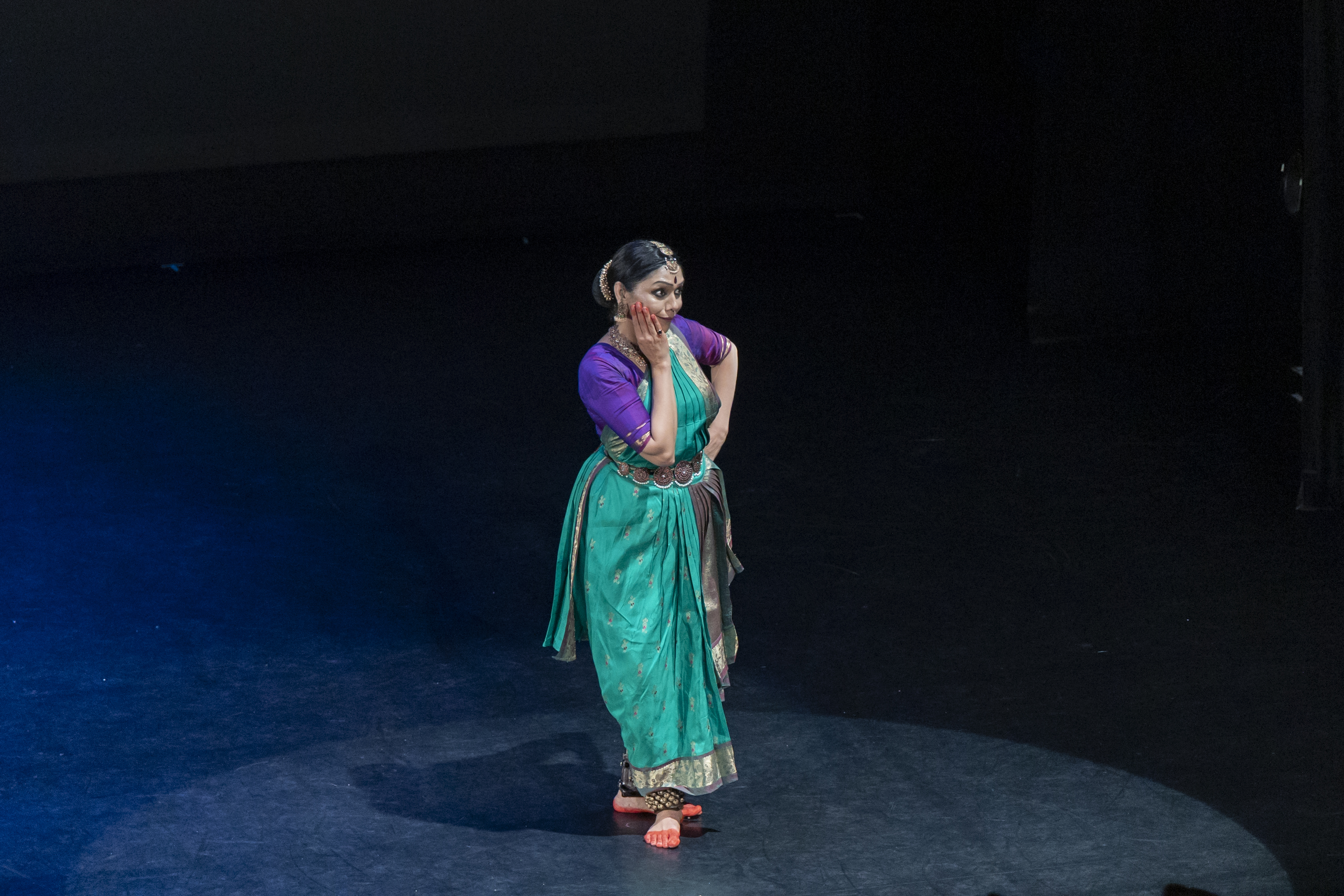 A standing woman in dressed in purple and turquoise lowers her gaze as her red-fingered hand brushes her cheek. ers her 