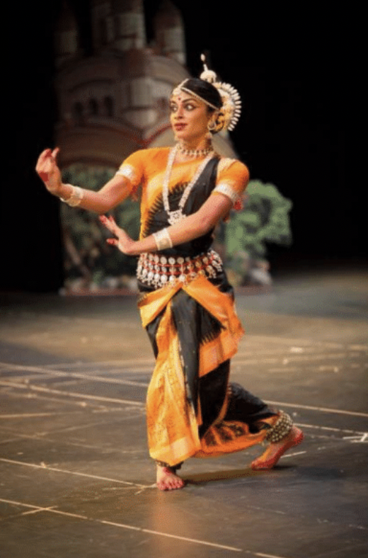 A woman dressed in yellow wearing an elaborate spikey headdress opens her eyes expressively wide. Her red-tipped hands are in front of her body as she stands on one leg with the other leg trailing behind.
