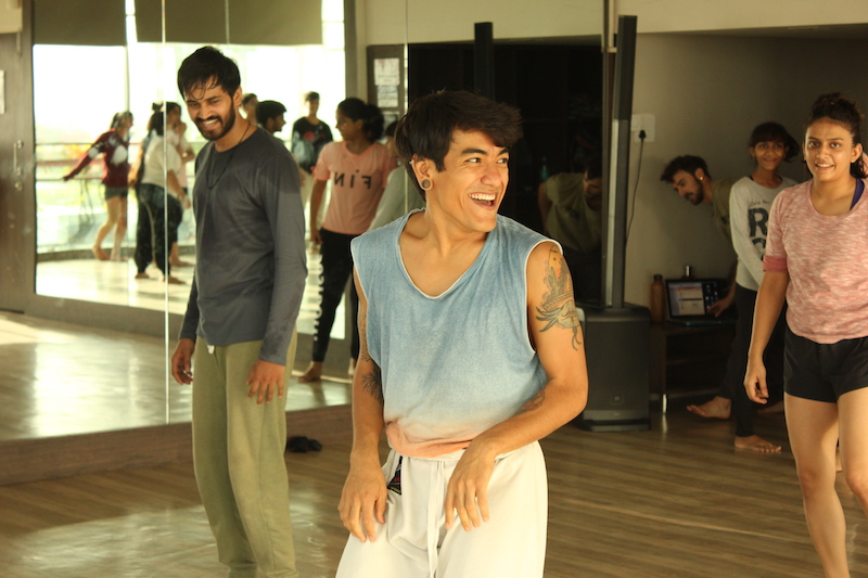 A dancer in a sleeveless tie dye shirt lead a group of dancers who are standing behind him in a dance studio