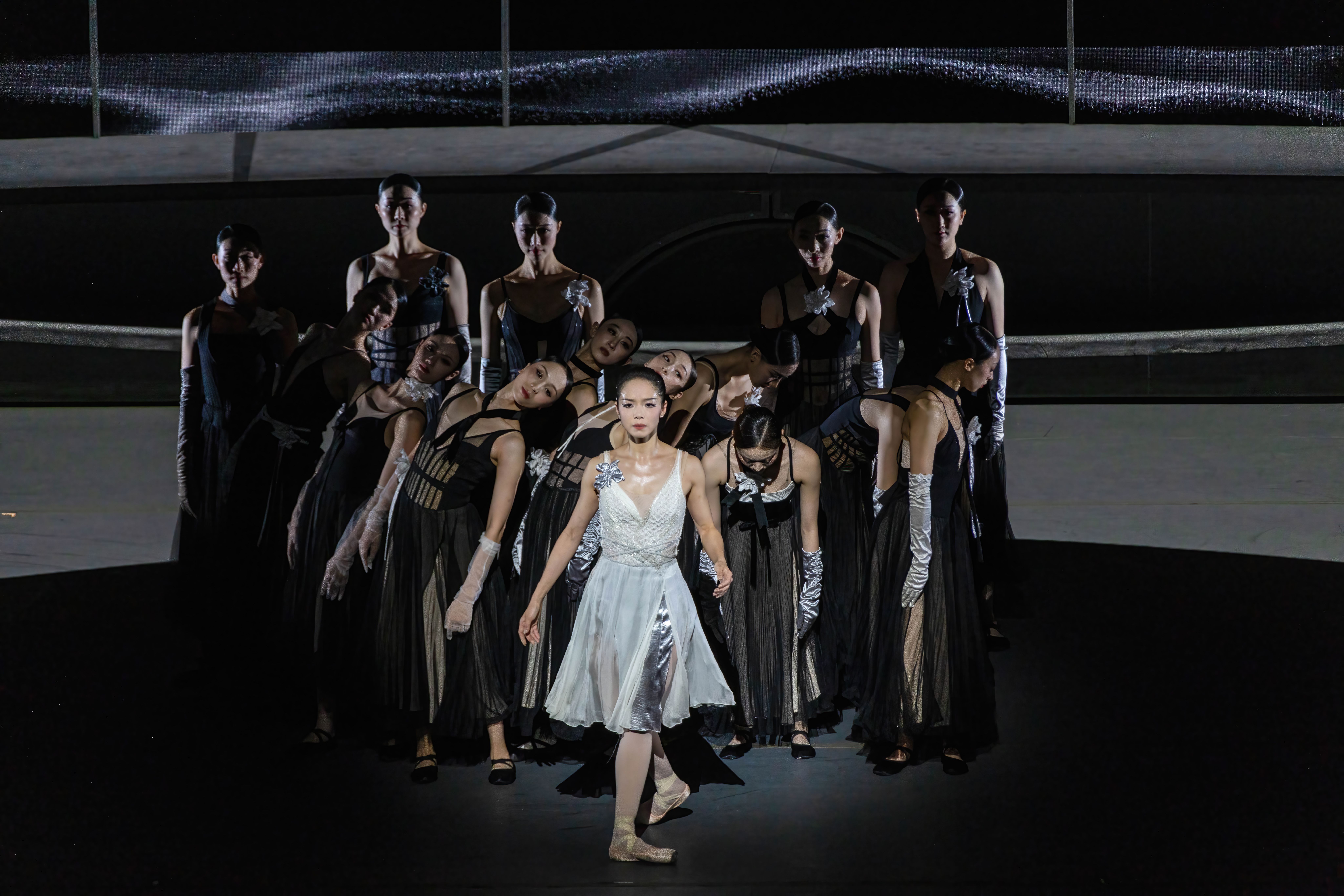 A woman costumed in a white dress faces the audience while leading a phalanx of women dancers dressed in black with silvery long opera gloves.