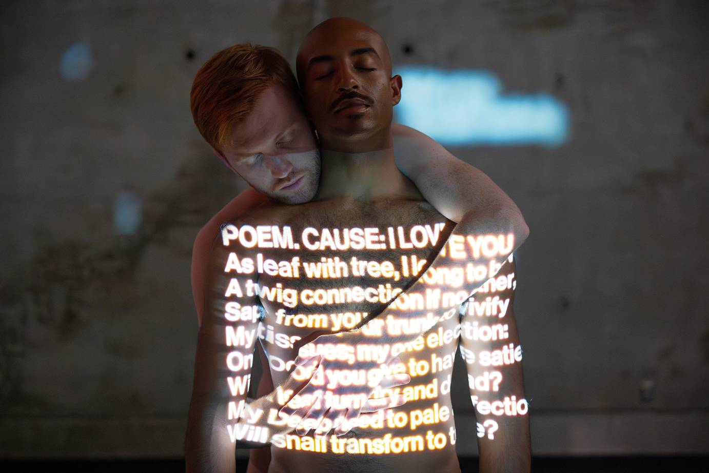 two men in an embrace, one behind the other, both have eyes closed as if in reverie, across their chests are words of love 