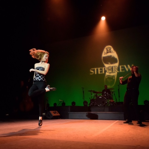 A woman step dancer hops in the air with one leg bent and crossed over her other thigh. A fiddler and drummer are in the background.