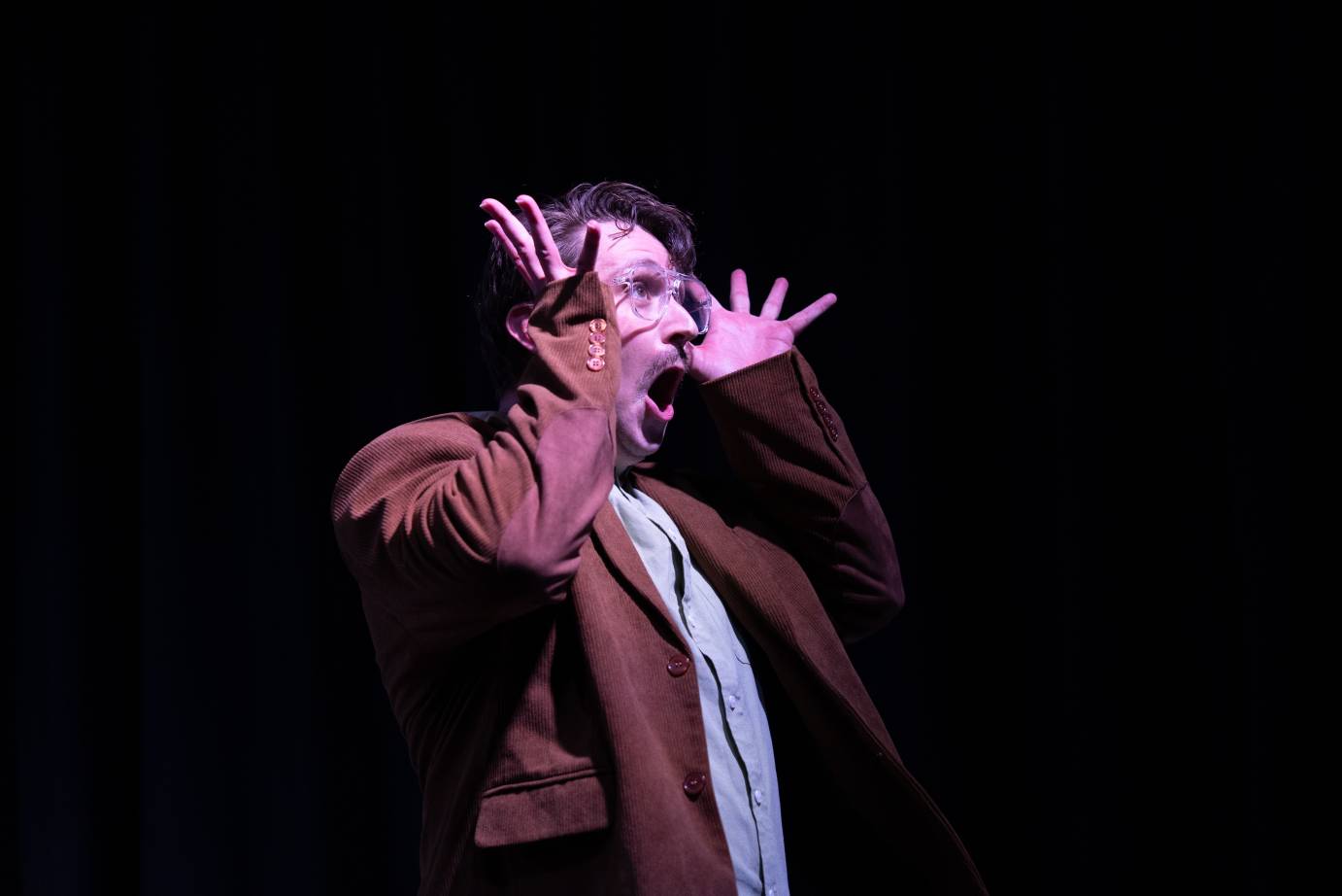 A bespeckled, mustachioed man, open-mouthed, opens his fingers wide on either side of his face.