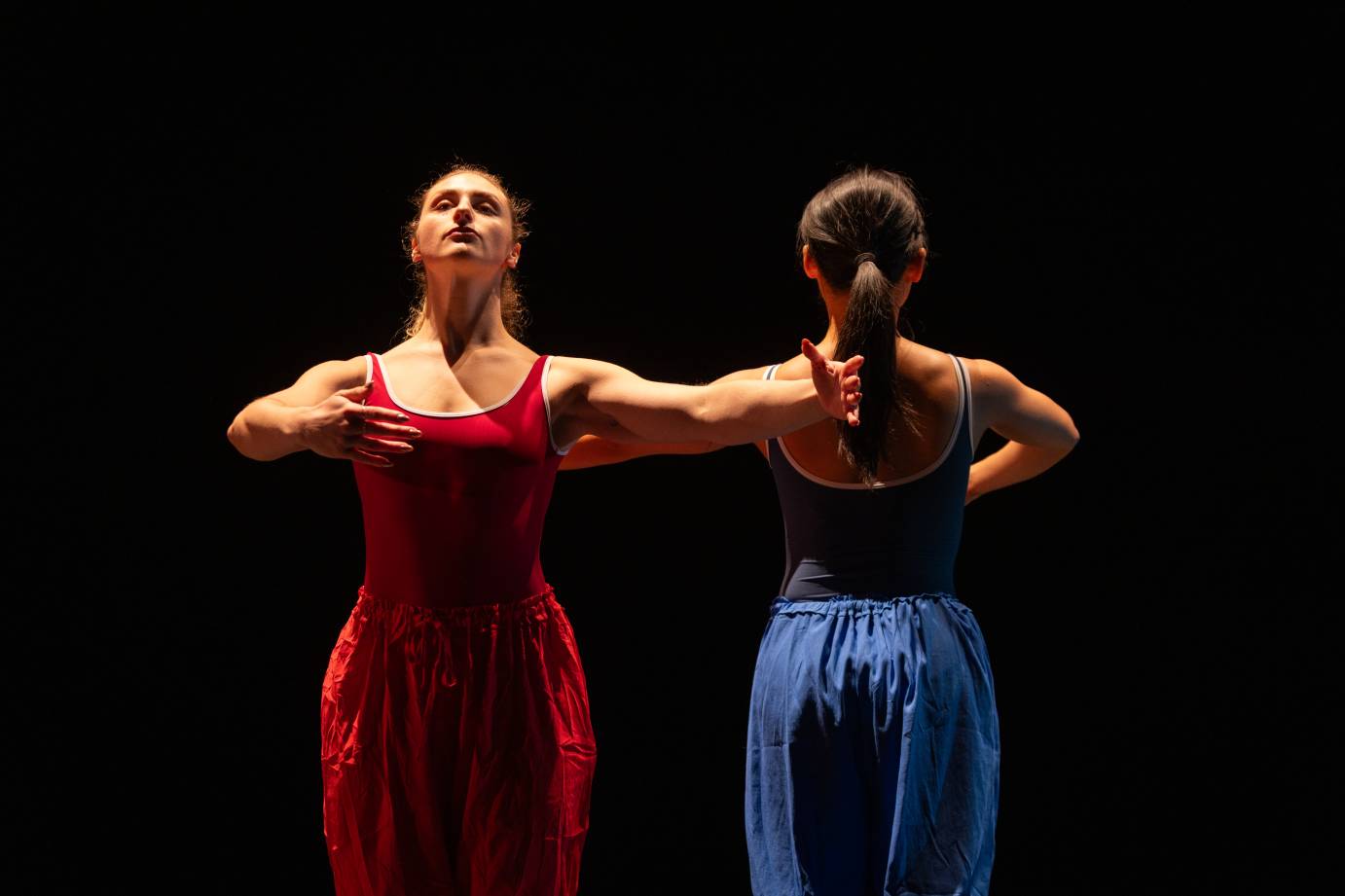 Standing side by side, one dancer in red and the other in blue, one facing upstage and the other downstage, curve their arms.