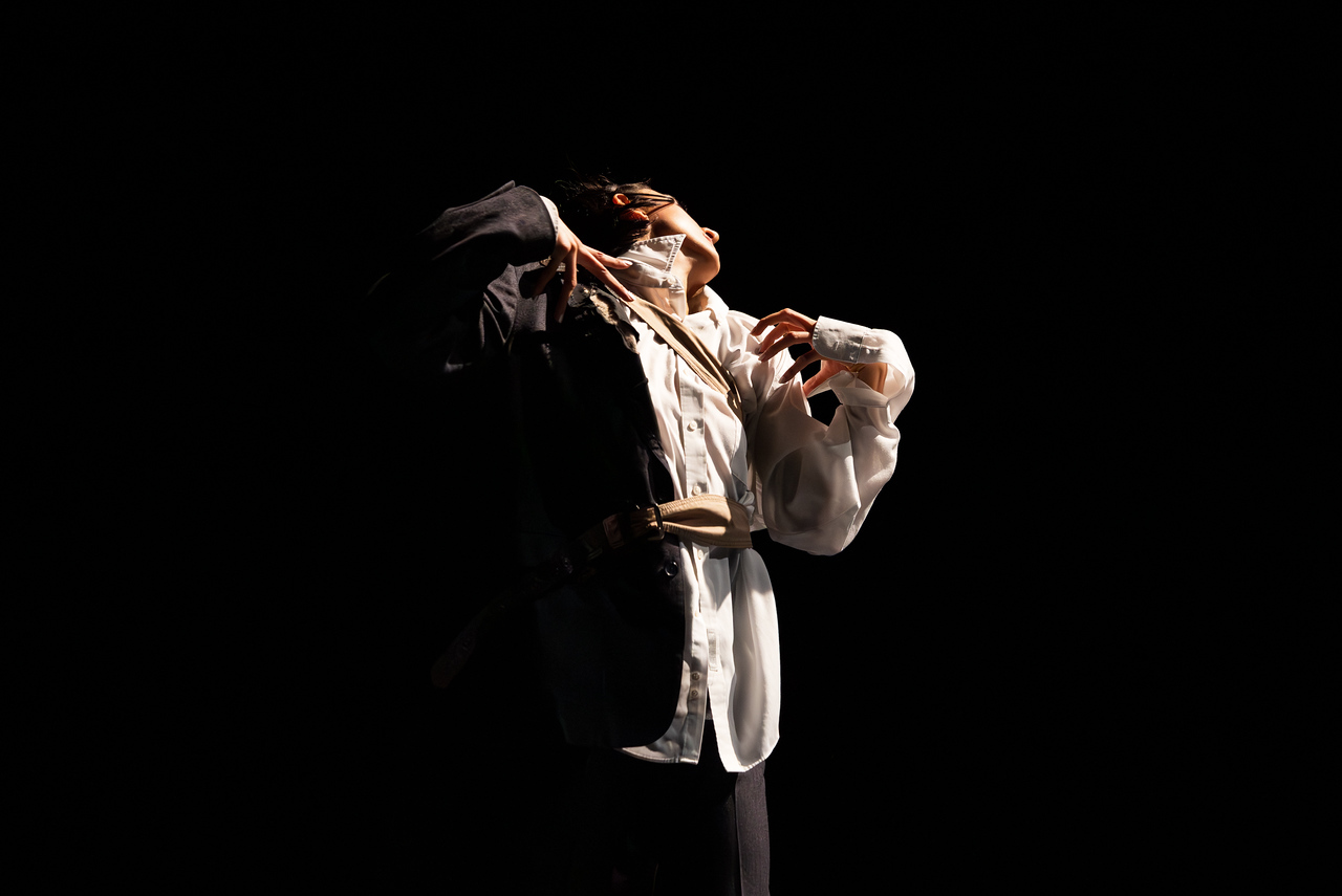 A dancer, spiraled body white shirt flying with splayed fingers is illuminated in half-light.