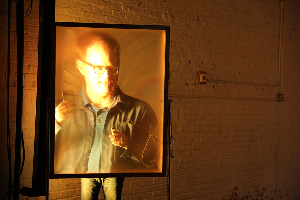 A man stands behind a glass screen with his image projected on it. It is hard to tell if this is a phtograph or live.