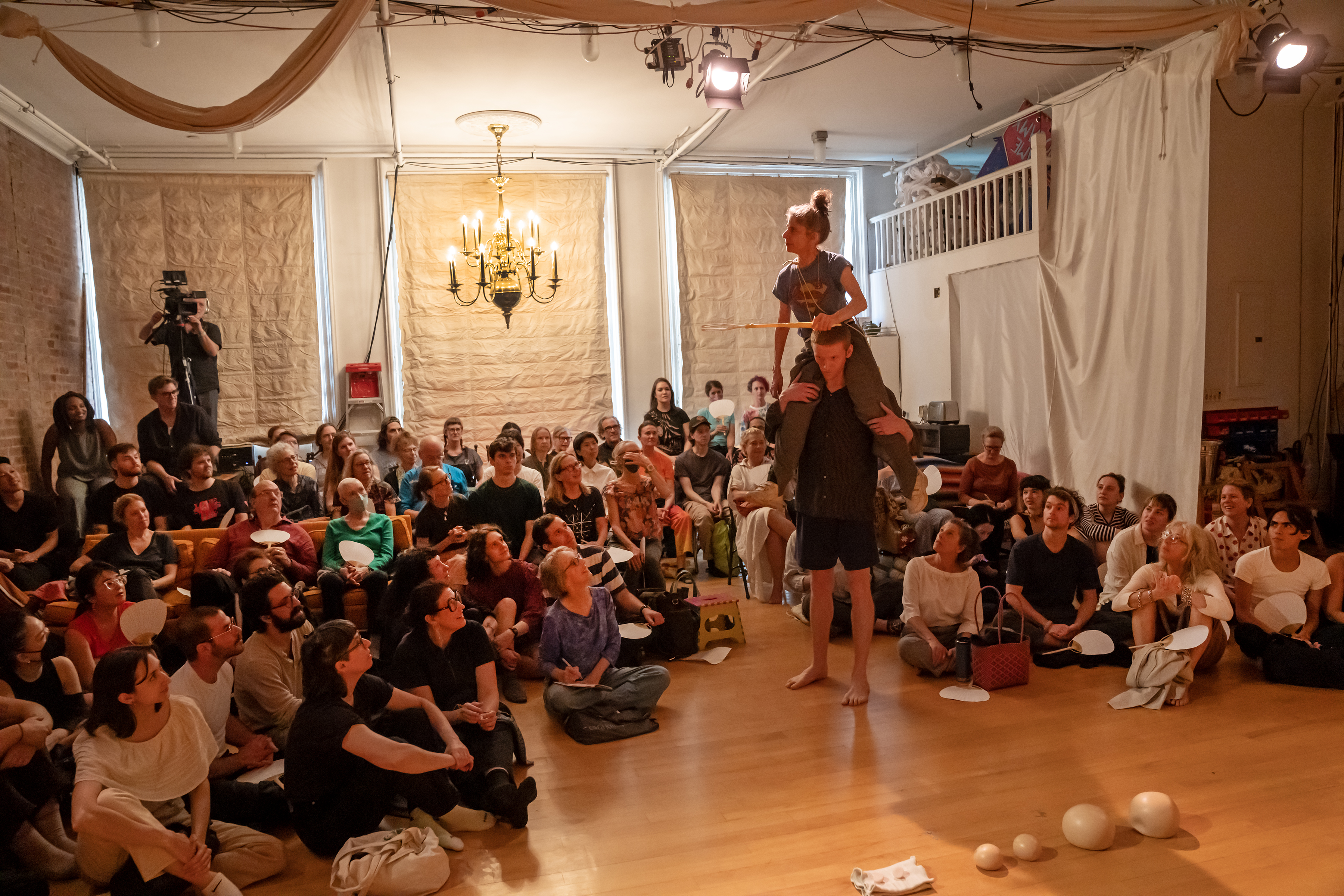 A loft space filled with audience on various chairs, sofas and the floor. A lit chandelier hangs above the crowd. A woman is perched on a tall man's back.