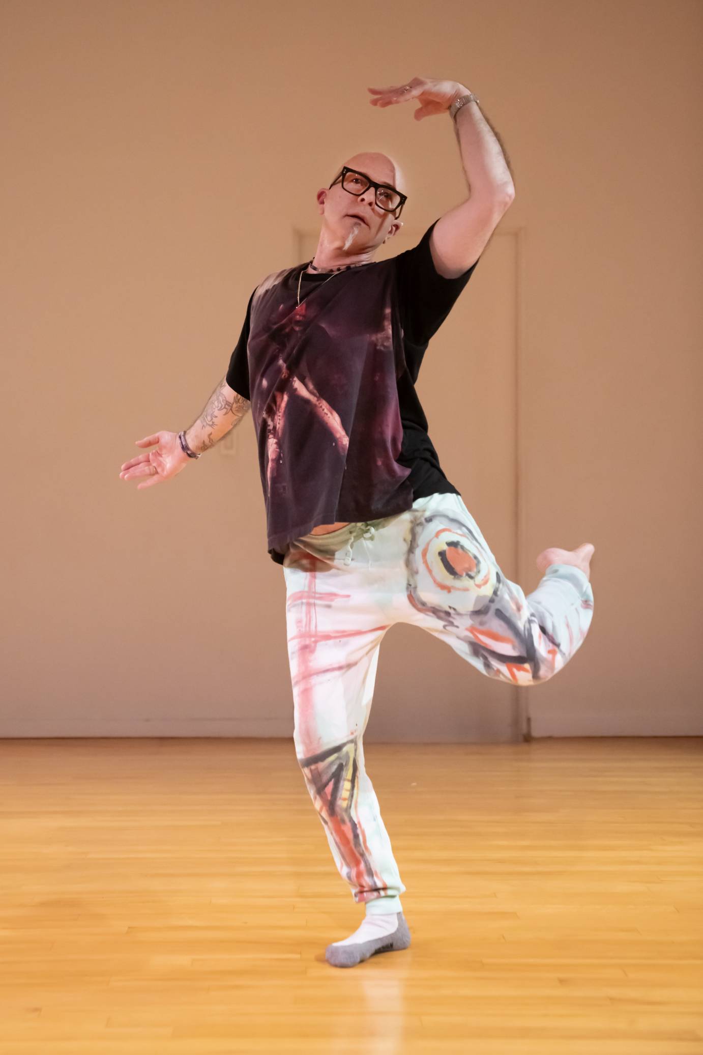 Man on releve, body bending to the left, dressed in fancifully-painted light sweatpants and black shirt, wearing large black-framed glasses, jewelry and facial hair.